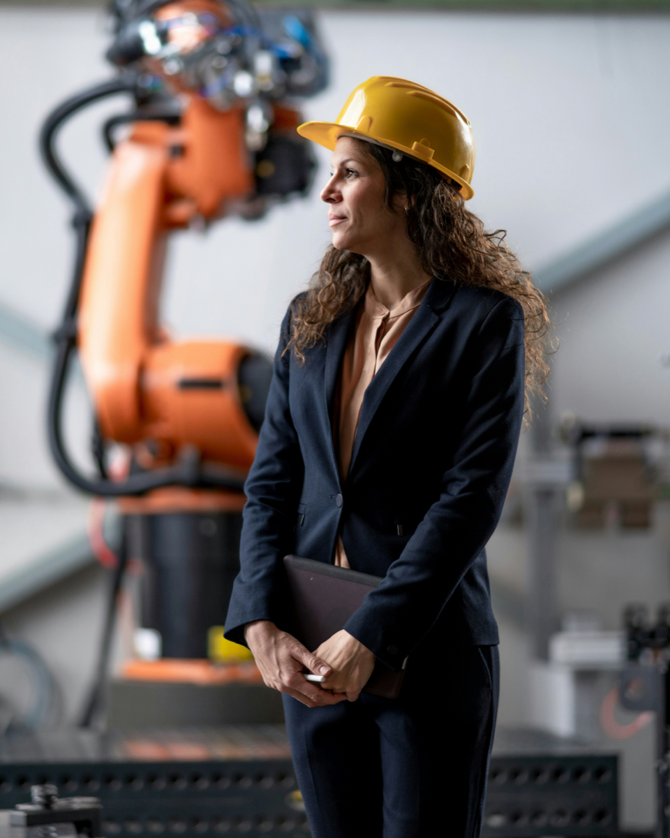 Businesswoman wearing a yellow safety helmet in a factory hall in front of an industrial robotic arm, considering heat recovery from corrosive flue gases.