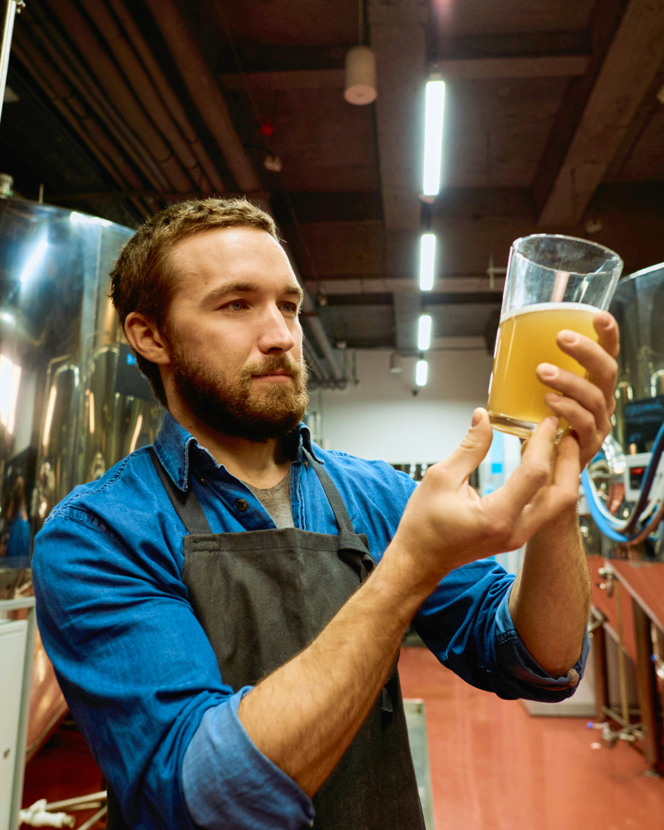 Brewer inspecting a glass of cloudy beer in a modern production facility, with shiny stainless steel tanks in the background.