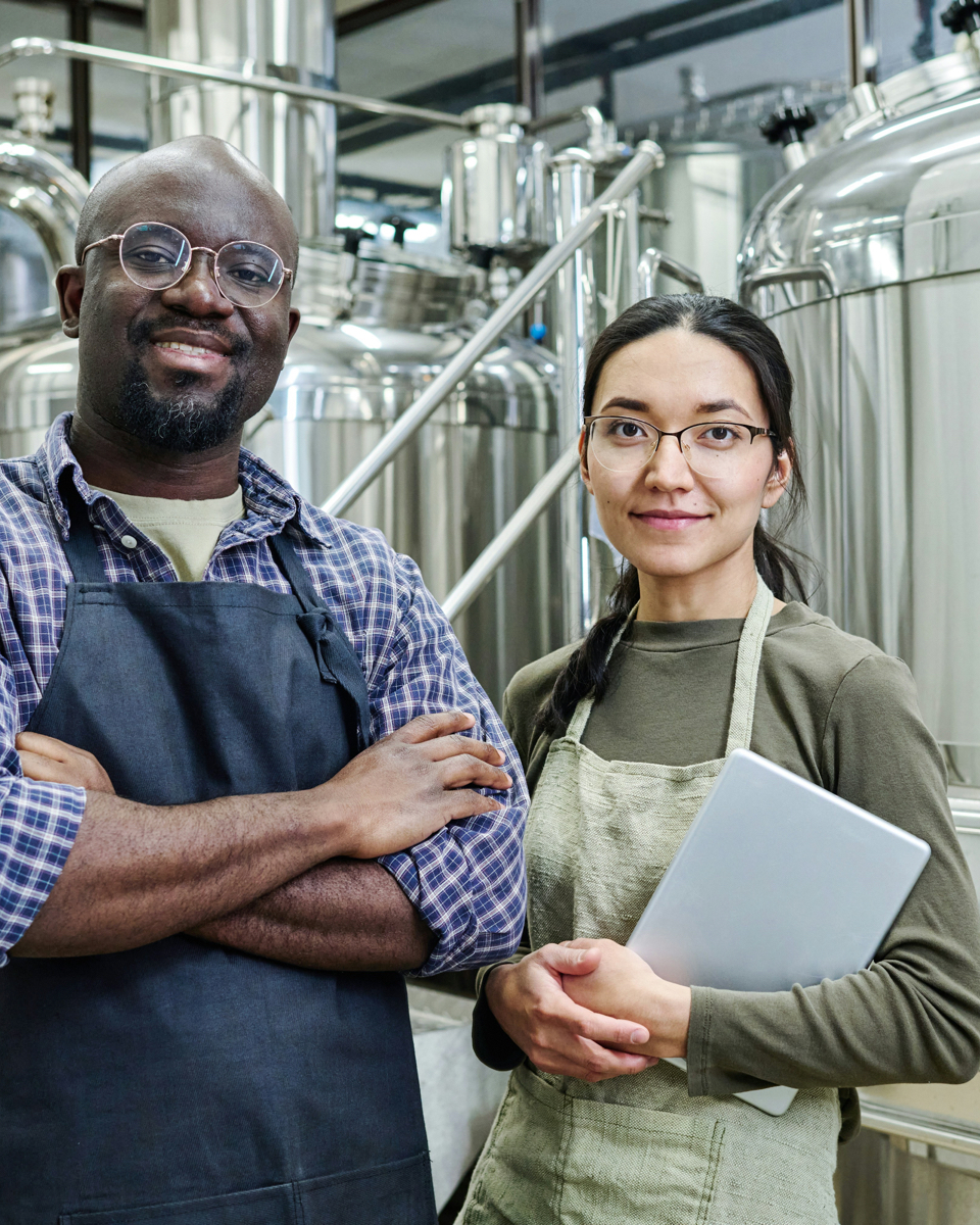 Two employees in an industrial facility with stainless steel tanks, wearing aprons and reviewing the results of the Heat Recovery Scan together.