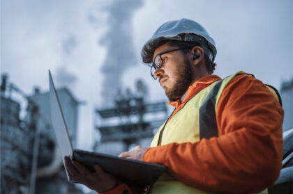 Industrial engineer with safety gear analyzing process data on a laptop in front of a factory chimney emitting flue gases.